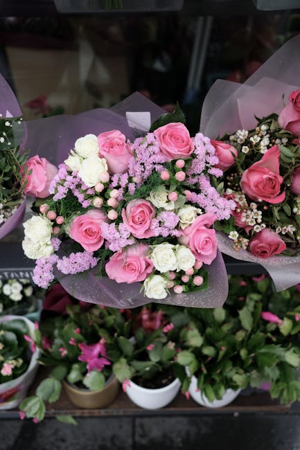 A floral bouquet featuring an arrangement of pink roses, white carnations, and light purple filler flowers, all beautifully wrapped in soft purple translucent paper with a subtle sheen. The bouquet is