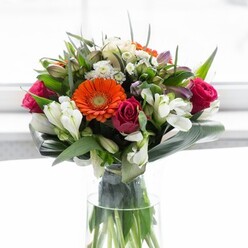Bathed in the gentle daylight that so often graces the leafy avenues of Knightsbridge, this lush bouquet tells a story of joy and sophistication. At its heart, a brilliant orange Gerbera daisy spreads its sunburst petals, beckoning the eye amidst a gathering of jewel-toned roses-some flushed deep with magenta, others unfurling burgundy and crimson. The interplay of pure white alstroemeria, with their striped petals, and shy, daisy-like chrysanthemums softens the arrangement, giving a cloud-like delicacy that tempers the more opulent blooms. Above, soft yellow and ivory roses offer a graceful balance, their presence mirrored by cool lavender sprigs peeking through. The whole composition is framed by luxurious green foliage-broad, glossy leaves that arc protectively, and spiky accents that add quiet energy. Resting in clear, fresh water, the bouquet looks as if it has been freshly gathered from a Knightsbridge market, its colours intensified by the diffused light pouring through a nearby window. This masterfully curated piece brings both comfort and celebration, making it an ideal choice for special occasions or to simply infuse a living space with the vibrancy and sophistication characteristic of the area.